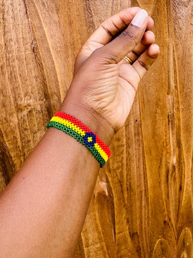 May include: A beaded bracelet featuring horizontal stripes of red, yellow, and green glass beads. The center of the bracelet displays a circular motif with a dark blue background, a yellow four-pointed star shape, and a green border.