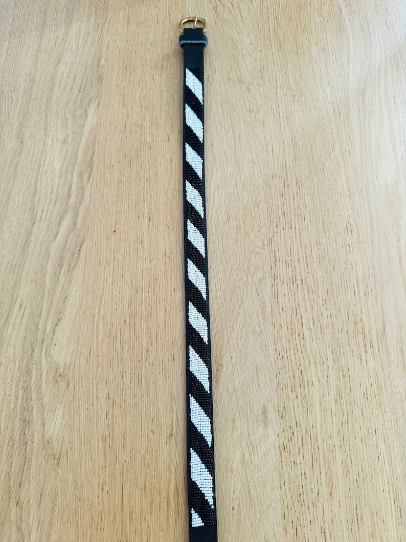 May include: A black leather belt with a white and black geometric pattern. The belt has a gold buckle.