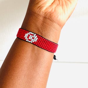 May include: A woven beaded bracelet featuring a vibrant red background with a prominent white circular symbol in the center. The bracelet is constructed from small, uniform beads arranged in a grid pattern, secured with black thread and a small black bead closure.
