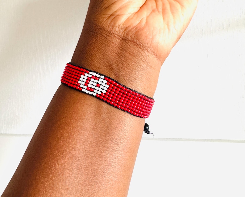 May include: A woven beaded bracelet featuring a vibrant red background with a prominent white circular symbol in the center. The bracelet is constructed from small, uniform beads arranged in a grid pattern, secured with black thread and a small black bead closure.
