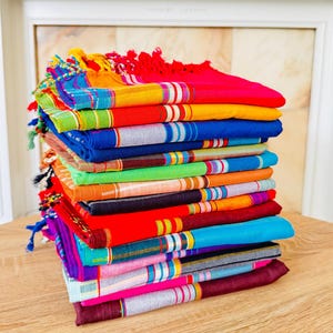 Può includere: Una pila di tessuti colorati tessuti con motivi a righe e bordi sfrangiati. I tessuti presentano una varietà di colori vivaci, tra cui rosso, blu, giallo e verde. I tessuti sono ben piegati e impilati l'uno sull'altro.
