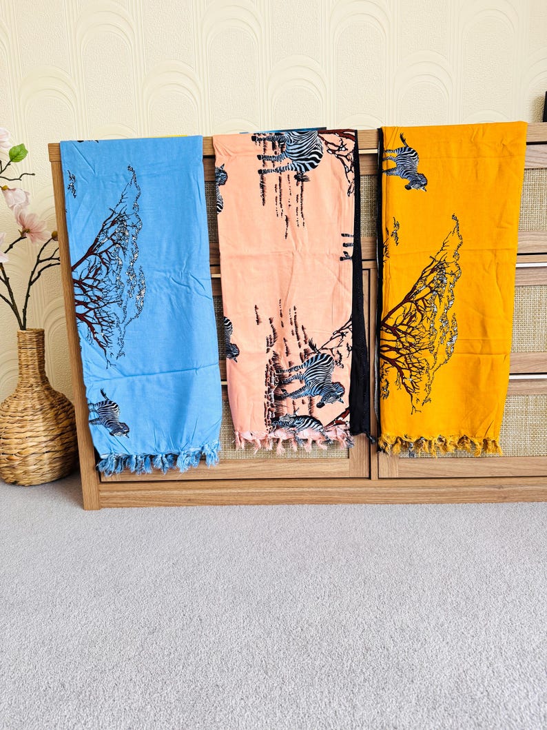 Pu&ograve; includere: Tre parei colorati appesi a un espositore in legno. I parei sono blu, pesca e gialli, ognuno con un motivo a stampa di albero e zebra. I bordi dei parei hanno nappe.