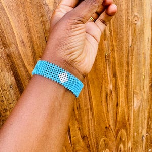 May include: A wide beaded bracelet featuring a repeating pattern of small, bright turquoise beads with a single, centered diamond-shaped motif composed of white beads. The bracelet is constructed from uniform, spherical beads arranged in a grid.