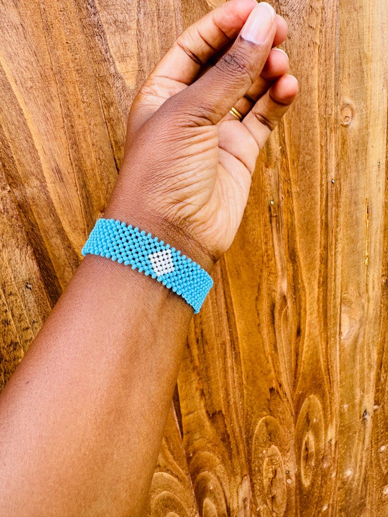 May include: A wide beaded bracelet featuring a repeating pattern of small, bright turquoise beads with a single, centered diamond-shaped motif composed of white beads. The bracelet is constructed from uniform, spherical beads arranged in a grid.