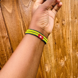 May include: A beaded bracelet featuring three horizontal rows of yellow beads separated by black bead lines. A rectangular red section is integrated into the design, displaying a small white cross symbol centered within the red beadwork.