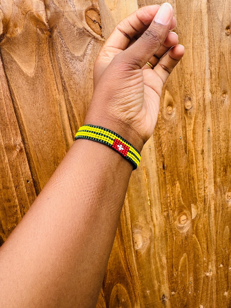 May include: A beaded bracelet featuring three horizontal rows of yellow beads separated by black bead lines. A rectangular red section is integrated into the design, displaying a small white cross symbol centered within the red beadwork.