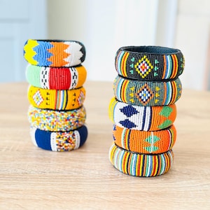 May include: Stacked beaded bracelets in various colors, including blue, orange, yellow, and green. The bracelets feature intricate patterns and designs, resting on a light-colored wooden surface. Each bracelet is approximately 1 inch wide.