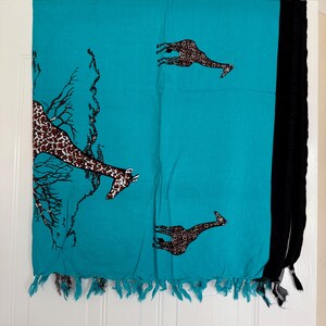 Pu&ograve; includere: Una sciarpa turchese con stampa di giraffe e frange nere. La sciarpa presenta giraffe marroni con un motivo a pois e un disegno ad albero. La sciarpa &egrave; realizzata in un materiale leggero.