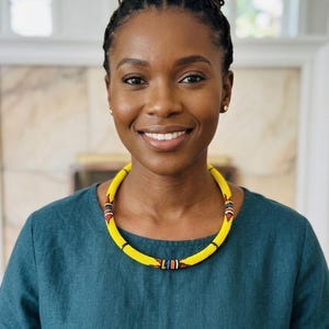 Op de afbeelding: Een vrouw draagt een gele kralenketting met rode, blauwe en witte accenten. De ketting is een choker-stijl en wordt gedragen over een teal groen topje. De vrouw heeft donker haar dat in een knot is opgestoken.