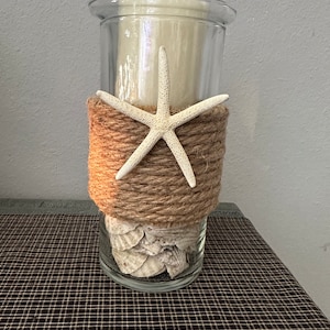 May include: A clear glass cylinder vase with a brown twine wrap and a white starfish. The vase is filled with white seashells and is sitting on a brown and white checkered surface.