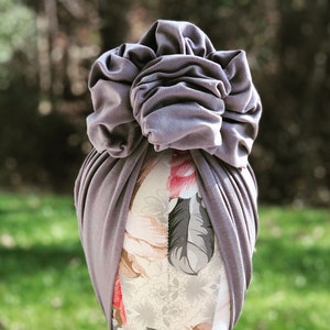 May include: A gray turban with a large, ruffled flower detail on top. The turban is wrapped around a mannequin head with a floral pattern. The background is blurred, showing green foliage.