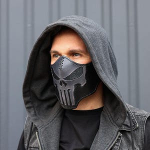 Peut inclure: Un masque facial en cuir noir avec un motif de cr&acirc;ne. Le masque est port&eacute; par une personne portant un sweat &agrave; capuche gris et un gilet en jean.