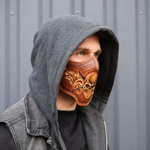 May include: A brown leather face mask with a skull design and intricate details. The mask is worn by a person with a gray hooded sweatshirt.