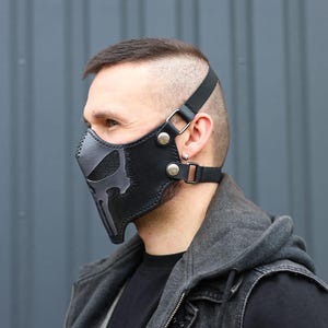 Peut inclure: Un masque facial en cuir noir avec un motif de cr&acirc;ne gris. Le masque est dot&eacute; de sangles r&eacute;glables et est port&eacute; par une personne.