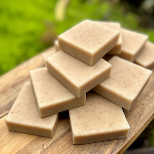 May include: A collection of square, light brown soaps with a speckled texture. The soaps are stacked on a wooden surface, with a blurred green background. The soaps appear to be handmade and are a natural colour.