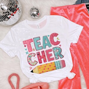May include: White t-shirt with the word "TEACHER" in colorful, patchwork-style letters and a yellow pencil graphic. The letters are in shades of pink, teal, and floral patterns. The shirt is paired with pink pants and disco balls.