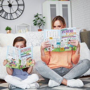 Pode incluir: Uma m&atilde;e e sua filha est&atilde;o sentadas no ch&atilde;o, cada uma lendo um livro colorido intitulado "Aprendiendo a Leer" com ilustra&ccedil;&otilde;es de crian&ccedil;as e letras. O livro tem uma capa azul com texto amarelo e uma imagem de desenho animado de crian&ccedil;as lendo.