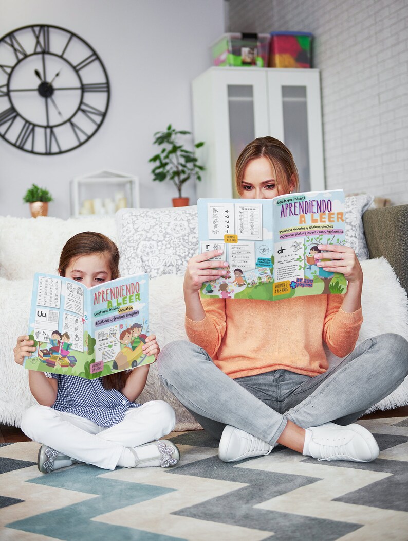 Pode incluir: Uma m&atilde;e e sua filha est&atilde;o sentadas no ch&atilde;o, cada uma lendo um livro colorido intitulado "Aprendiendo a Leer" com ilustra&ccedil;&otilde;es de crian&ccedil;as e letras. O livro tem uma capa azul com texto amarelo e uma imagem de desenho animado de crian&ccedil;as lendo.