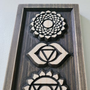 May include: A wooden incense holder with seven carved chakra symbols. The symbols are in a vertical row, starting with the crown chakra at the top and ending with the root chakra at the bottom. The holder is stained a dark brown color.