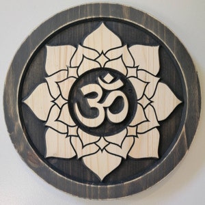 May include: A wooden wall hanging with a carved Om symbol surrounded by a lotus flower design. The background is dark brown and the flower and symbol are light wood.