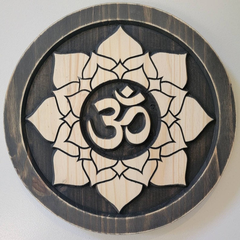 May include: A wooden wall hanging with a carved Om symbol surrounded by a lotus flower design. The background is dark brown and the flower and symbol are light wood.