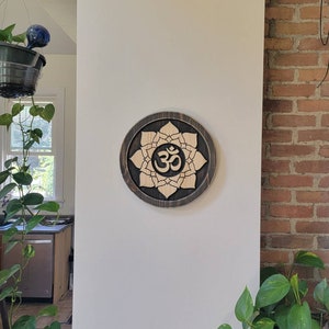 May include: A wooden wall hanging with a dark brown background and a light brown lotus flower design. The center of the flower features the Om symbol in a darker brown color.