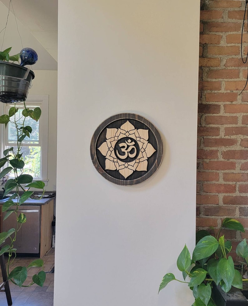 May include: A wooden wall hanging with a dark brown background and a light brown lotus flower design. The center of the flower features the Om symbol in a darker brown color.