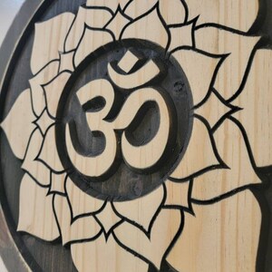 May include: A wooden wall hanging with a black and white carved Om symbol surrounded by a lotus flower design.