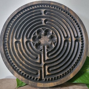 Puede incluir: Un laberinto circular de madera oscura con un diseño de flor tallado en el centro. El laberinto está rodeado por un borde de cuentas.