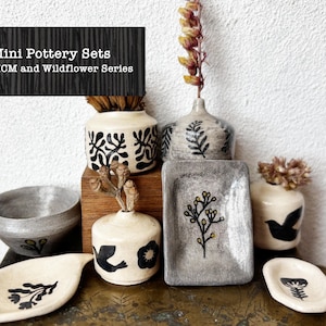May include: A selection of miniature pottery sets, including bowls, vases, and trays. The pieces are in neutral tones with black floral and geometric designs. Some pieces hold dried flowers. The text "Mini Pottery Sets MCM and Wildflower Series" is at the top.
