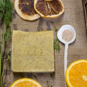 Pode incluir: Uma barra de sabonete natural de cor verde e amarela. O sabonete tem as palavras "Bath & Body" em relevo. O sabonete está rodeado de fatias de laranja, cenouras e paus de canela.