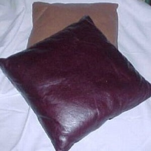 Könnte beinhalten: Zwei dekorative Kissen, eines braun und eines burgunderrot, liegen auf einer weißen Oberfläche. Das burgunderrote Kissen ist aus einem lederartigen Material gefertigt.