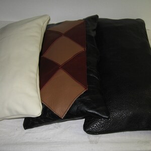 May include: Three rectangular leather pillows in cream, brown and black. The center pillow has a diamond pattern in shades of brown and burgundy. The black pillow has a textured surface. The pillows are arranged side by side.