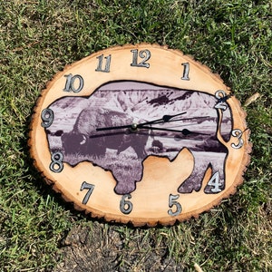 May include: A wooden clock with a bison silhouette cut out. The clock face is a black and white photo of a bison in a field. The numbers 1 through 12 are printed on the wood around the edge of the clock.