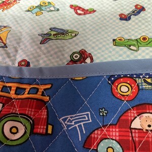 May include: A quilted fabric with a blue border and a pattern of cartoon vehicles. The top layer has a light blue gingham pattern with airplanes and cars. The bottom layer is blue with a diamond pattern and images of a fire truck and a car.