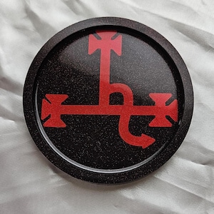 May include: Round black coaster with a red symbol. The coaster has a glittery texture and a raised edge. The red symbol is a stylized design with a cross-like shape at the top and sides, and a curved line with an arrow.