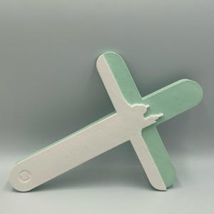 Puede incluir: Una cruz bicolor en blanco y verde menta. La cruz presenta una silueta detallada de una persona con los brazos extendidos. El diseño es simple y moderno, con un acabado mate.