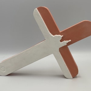 Puede incluir: Una escultura en forma de cruz de dos tonos. La parte delantera es blanca y la trasera es de color marrón rojizo. Una silueta blanca de una figura está recortada en la parte delantera. La escultura está hecha de un material sólido.