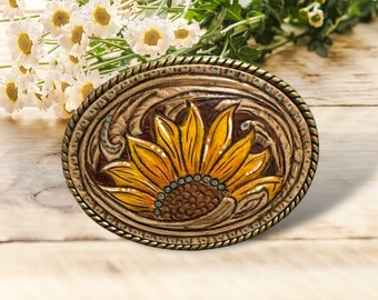 May include: A brown leather belt buckle featuring a sunflower design. The sunflower is yellow with a brown centre and turquoise accents.