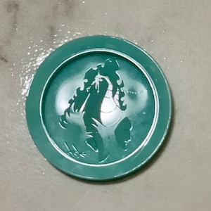 May include: A teal green round coaster with a white silhouette of a dragon in the center.