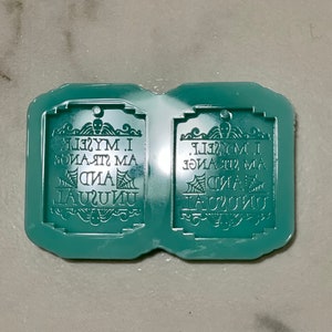May include: Two teal blue silicone molds for making resin charms. Each mold features a gothic-style design with the text "I am strange and unusual myself."