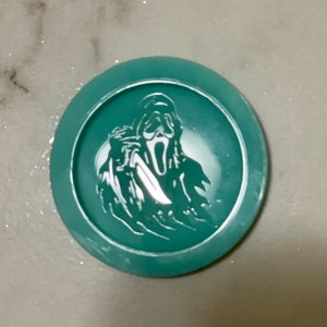 May include: A teal round coaster with a white silhouette of a ghost holding a knife. The ghost is wearing a mask with a scream face.