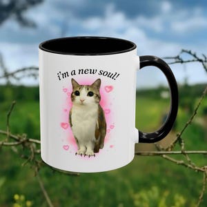 May include: White ceramic mug with a black interior and handle. The mug features a cat illustration surrounded by pink hearts and the text "i'm a new soul!". The background is a blurred outdoor scene.