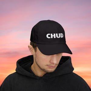 May include: Black baseball cap with the white embroidered word "CHUD" across the front. The cap has a curved brim and is worn by a person. The background is a gradient of pink, orange, and yellow.