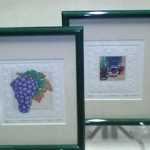 May include: Two framed prints with green frames. The left print features a cluster of purple grapes with green leaves. The right print features a bottle of wine and two glasses of red wine. Both prints have white mats and white backgrounds.