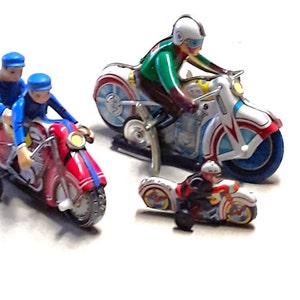 以下が含まれることがあります： ライダー付きのヴィンテージなブリキのおもちゃのオートバイ3台。オートバイは赤、白、青で、黒のアクセントが付けられています。ライダーはヘルメットをかぶっており、さまざまなポーズをとっています。