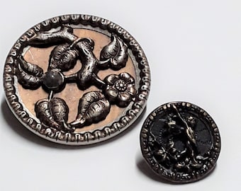 May include: Two antique metal buttons with floral designs. The larger button is silver with a copper center and features a raised floral design. The smaller button is bronze with a raised floral design.