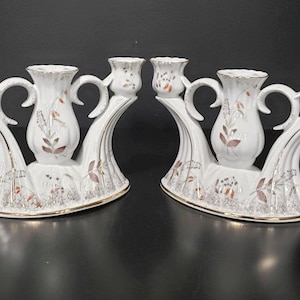 May include: A pair of white porcelain candle holders with gold trim and floral designs. Each holder has three candle cups and a curved base.