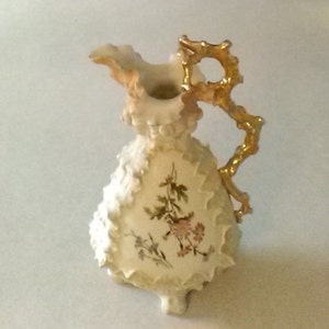 May include: A white porcelain pitcher with a gold handle and floral design. The pitcher has a scalloped edge and a delicate floral pattern.