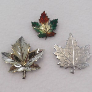 May include: Three maple leaf brooches. One is silver, one is silver and gold, and the third is painted with red, green, and gold. The brooches are likely made of metal and are displayed on a white surface.